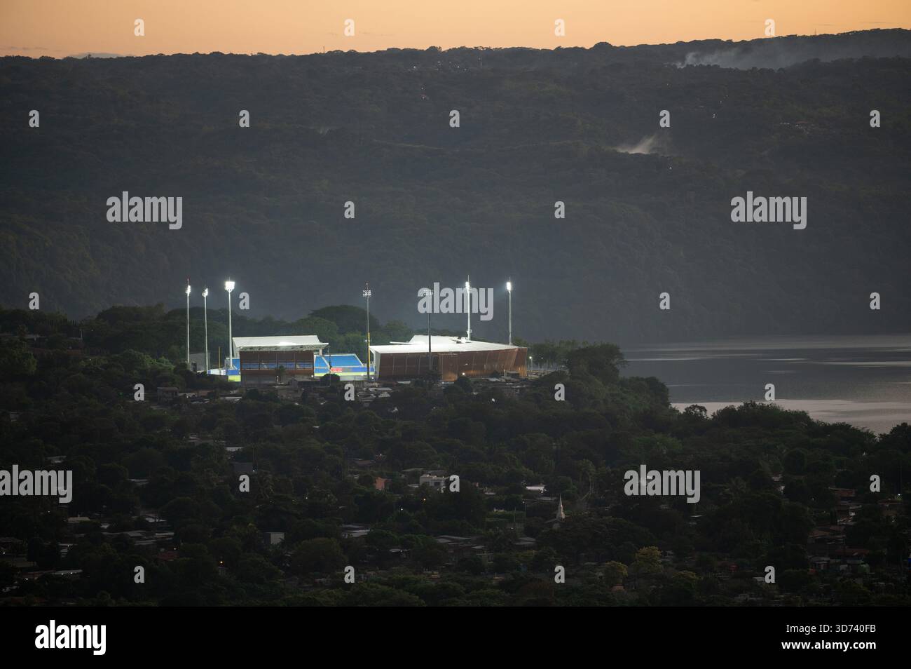 Helle Lichter erstrahlen auf einem Sportkomplex vor einer wunderschönen Abendkulisse mit weit entfernten Hügeln und einem See in der Nähe. Stockfoto