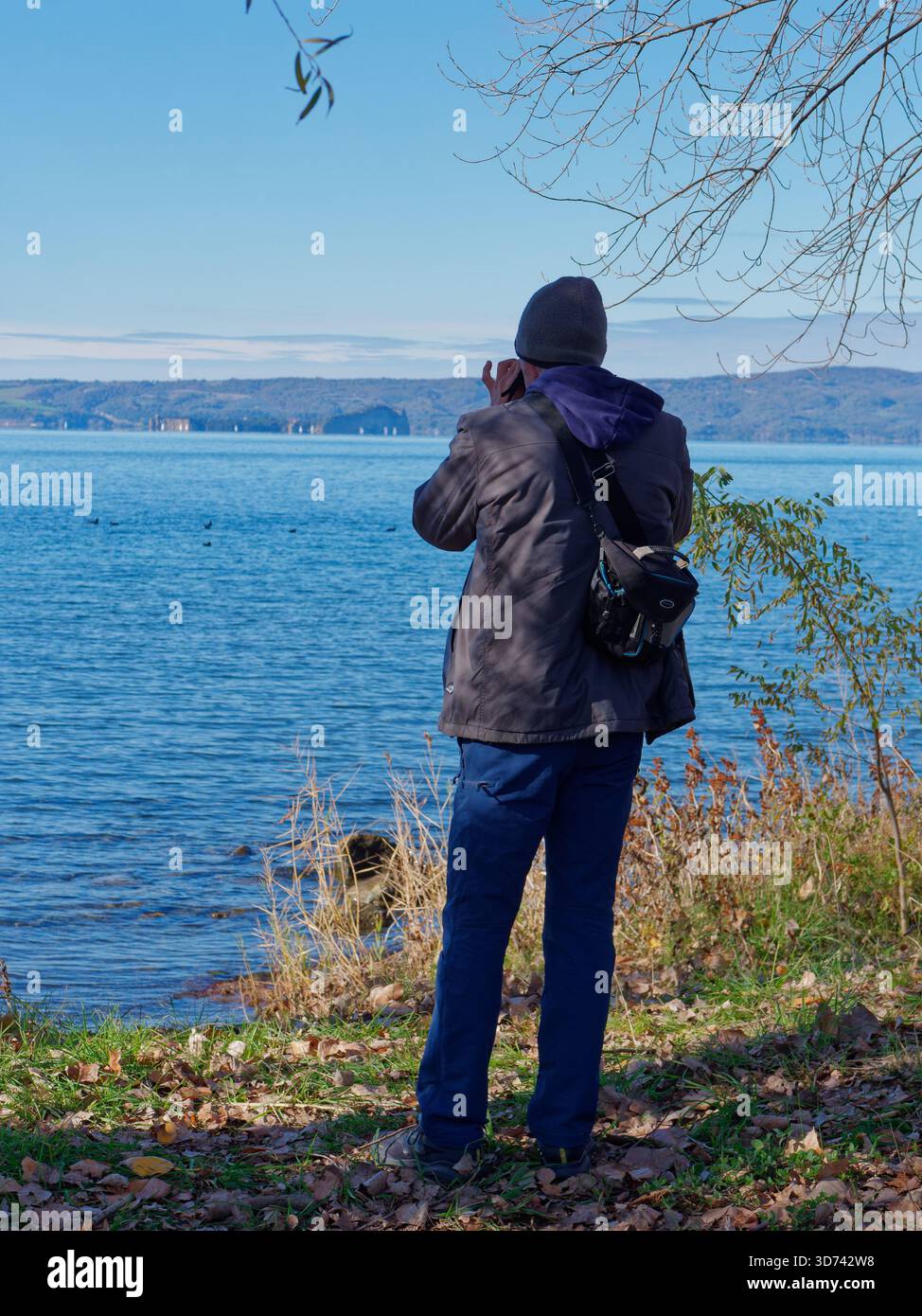 Mann mit Wollmütze macht ein Foto vom Bolsena-See aus Montefiascone, Latium, Italien. November 2025 Stockfoto
