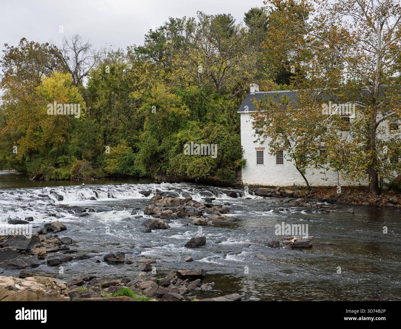 Ein großer Blick auf ein weißes historisches Mühlengebäude neben dem Brandywine Creek mit rauschenden Stromschnellen und Felsen im Vordergrund. Stockfoto