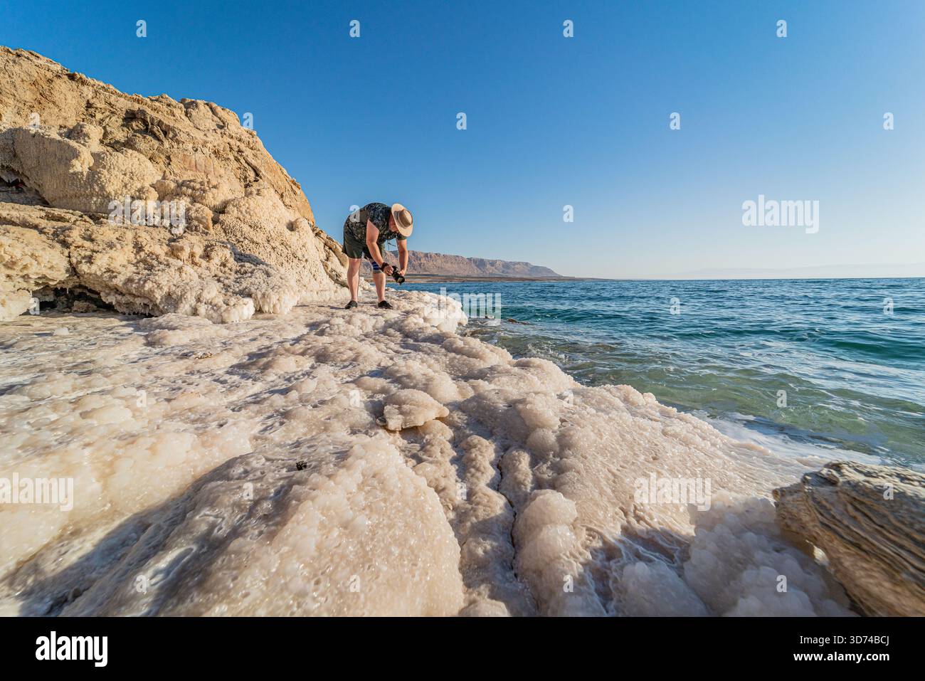 Fotograf, der die Salzküste des Toten Meeres, die weiße Mineralkruste unter hellem Sonnenlicht, das ruhige türkisfarbene Wasser in einer beeindruckenden natürlichen Landschaft einfängt. Stockfoto