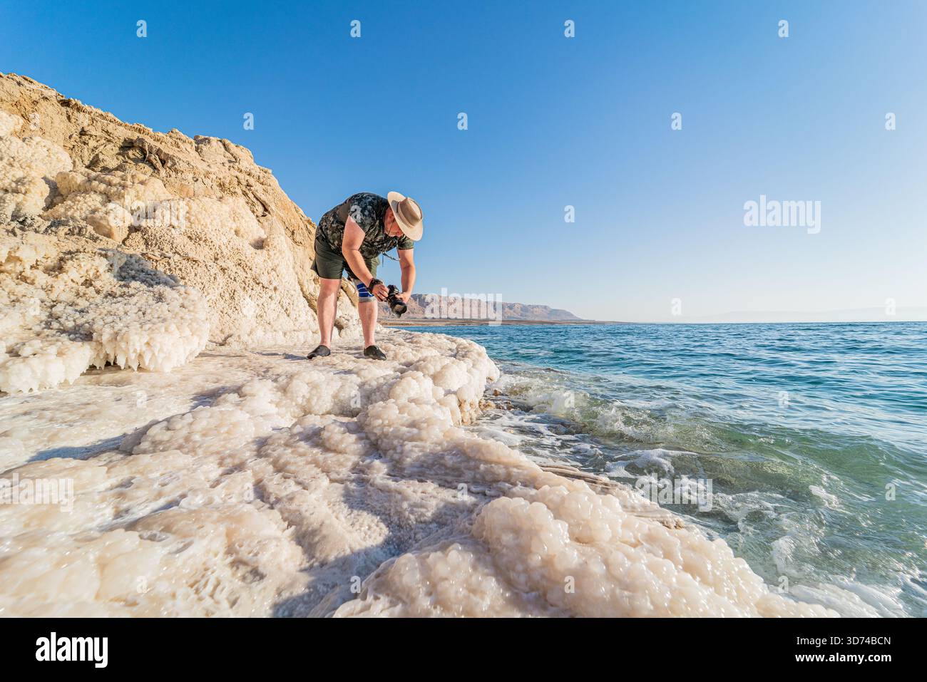 Fotograf, der die Salzküste des Toten Meeres, die weiße Mineralkruste unter hellem Sonnenlicht, das ruhige türkisfarbene Wasser in einer beeindruckenden natürlichen Landschaft einfängt. Stockfoto