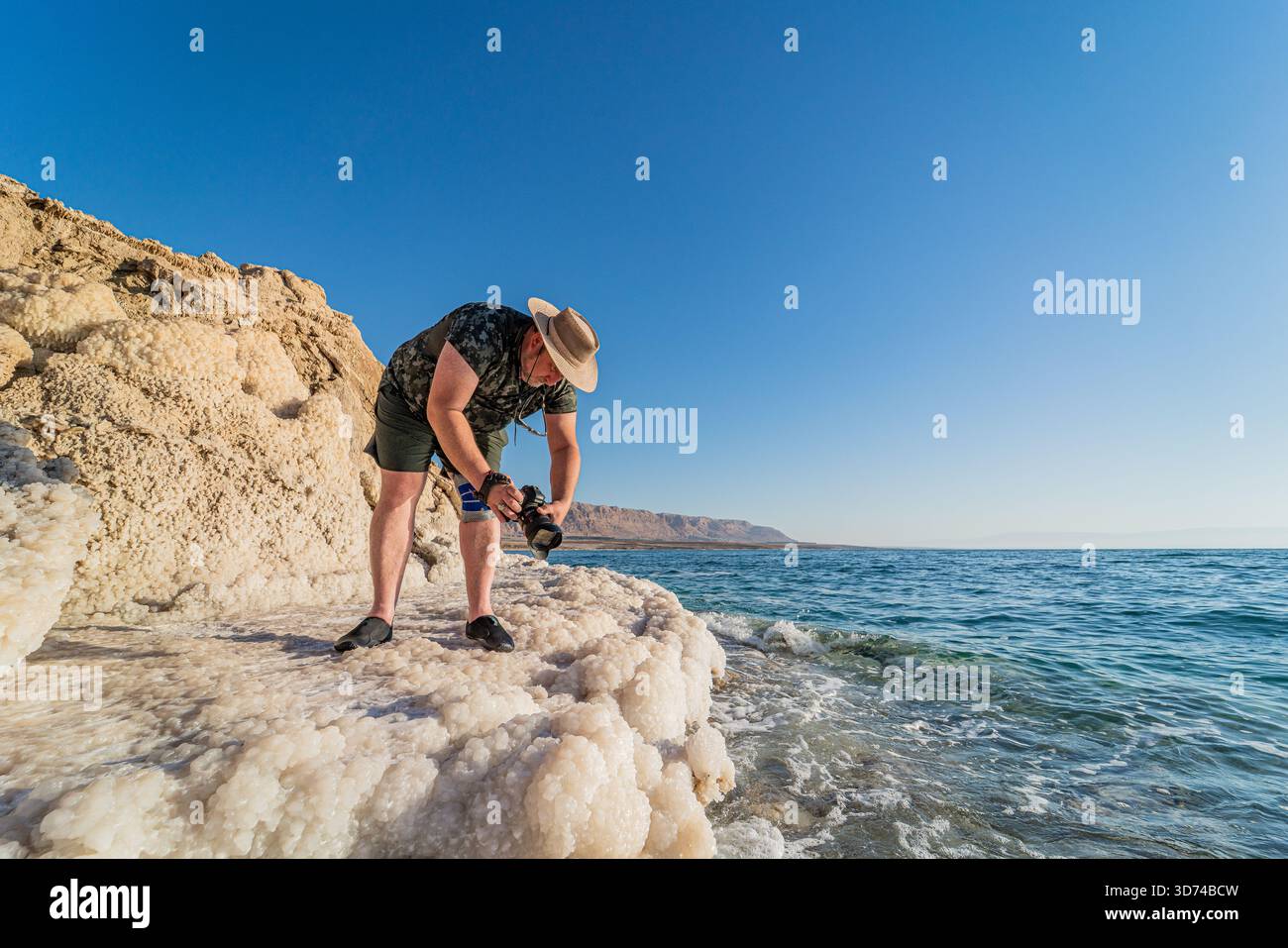 Fotograf, der die Salzküste des Toten Meeres, die weiße Mineralkruste unter hellem Sonnenlicht, das ruhige türkisfarbene Wasser in einer beeindruckenden natürlichen Landschaft einfängt. Stockfoto