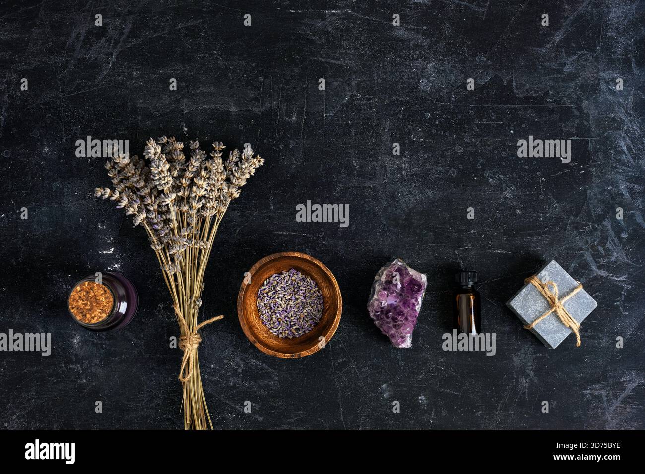 Getrocknete Lavendelblüten, Holzschale gefüllt mit Lavendel, Amethyst-Kristall, Flasche mit ätherischem Öl und handgemachter Seife auf strukturiertem schwarzem Hintergrund. Stockfoto