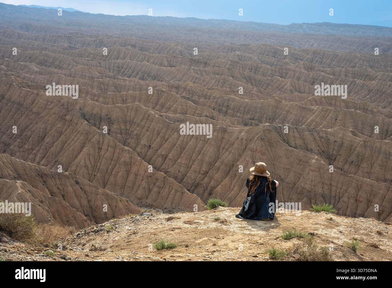 Allein Reisender mit Hut und Rucksack sitzt am Rand einer hohen Klippe und blickt über die weitläufige, tief erodierte, strukturierte Landschaft der Ak- Stockfoto