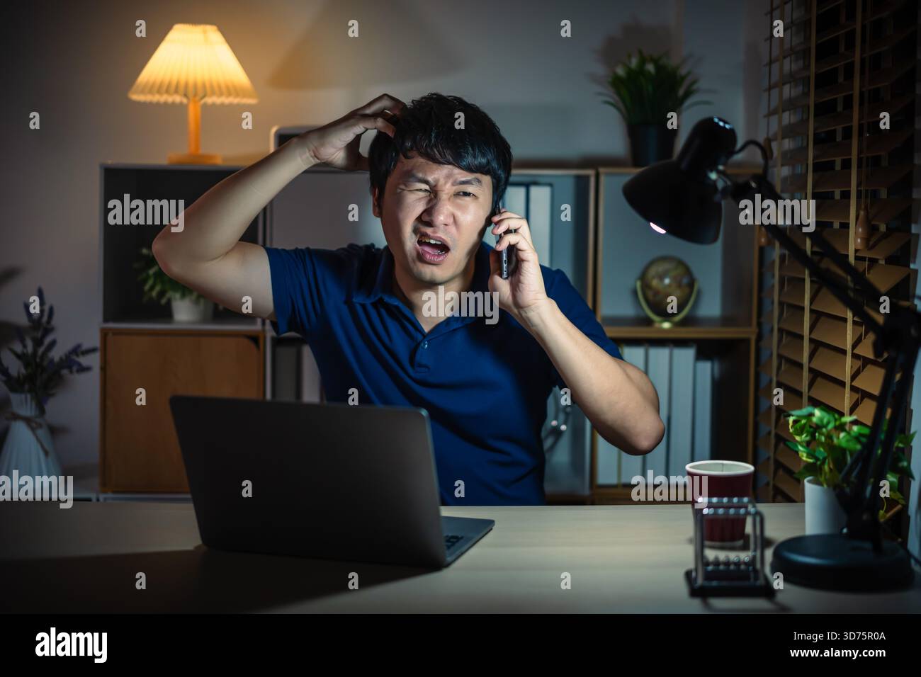 Gestresster Mann, der nachts mit dem Smartphone spricht, während er mit einem Laptop arbeitet Stockfoto