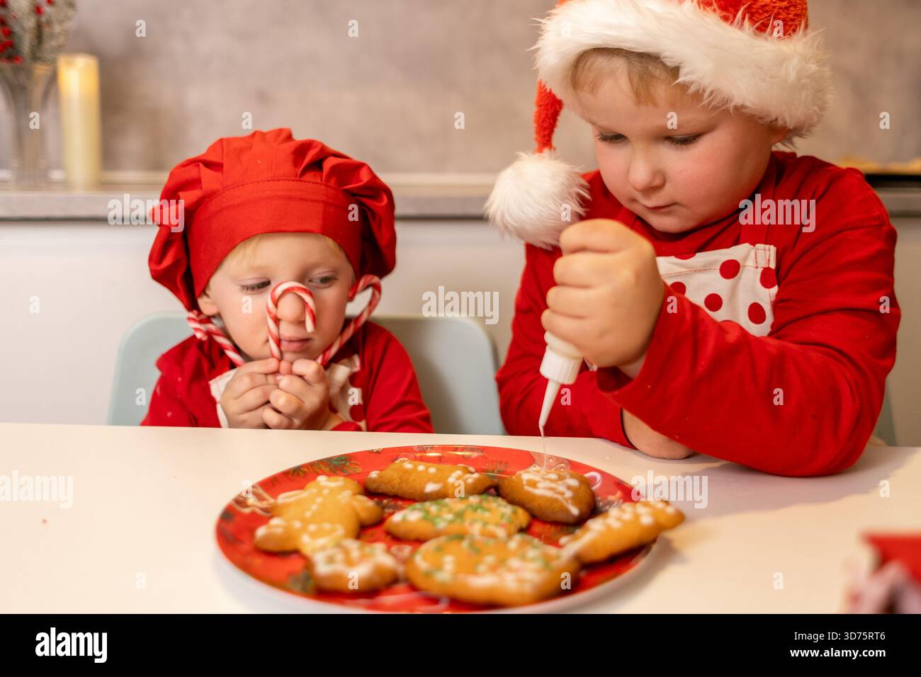 Zwei Kinder in Weihnachtskleidung schmücken Kekse mit Glasur. Ein Kind hält einen Zuckerrohr, während das andere den Keksen auf dem vorsichtig eine Glasur verleiht Stockfoto