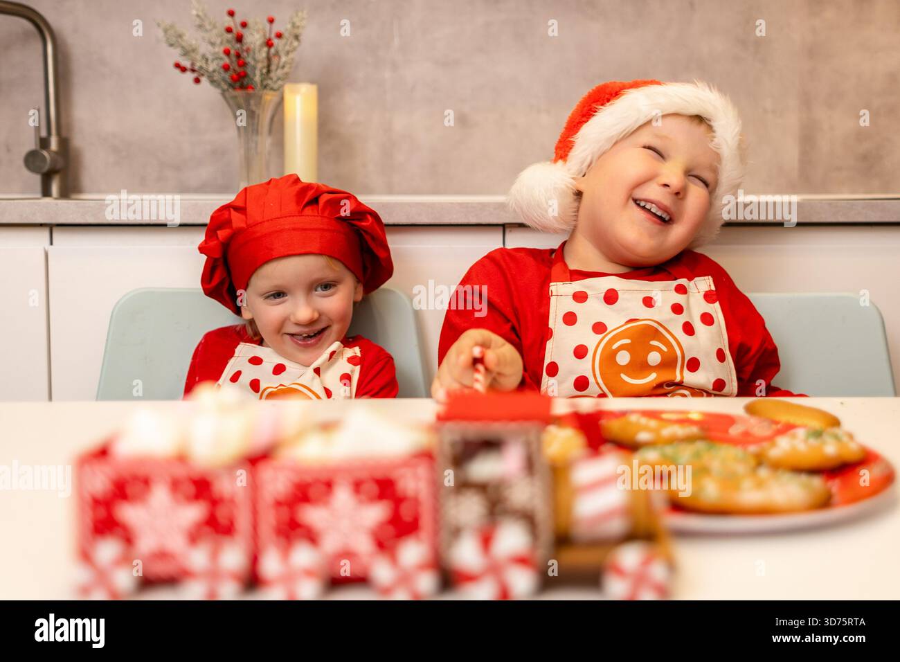 Zwei fröhliche Kinder in festlichen Outfits lachen, während sie an einem Tisch mit dekorierten Keksen in einer gemütlichen Küche sitzen. Sie feiern die Stockfoto