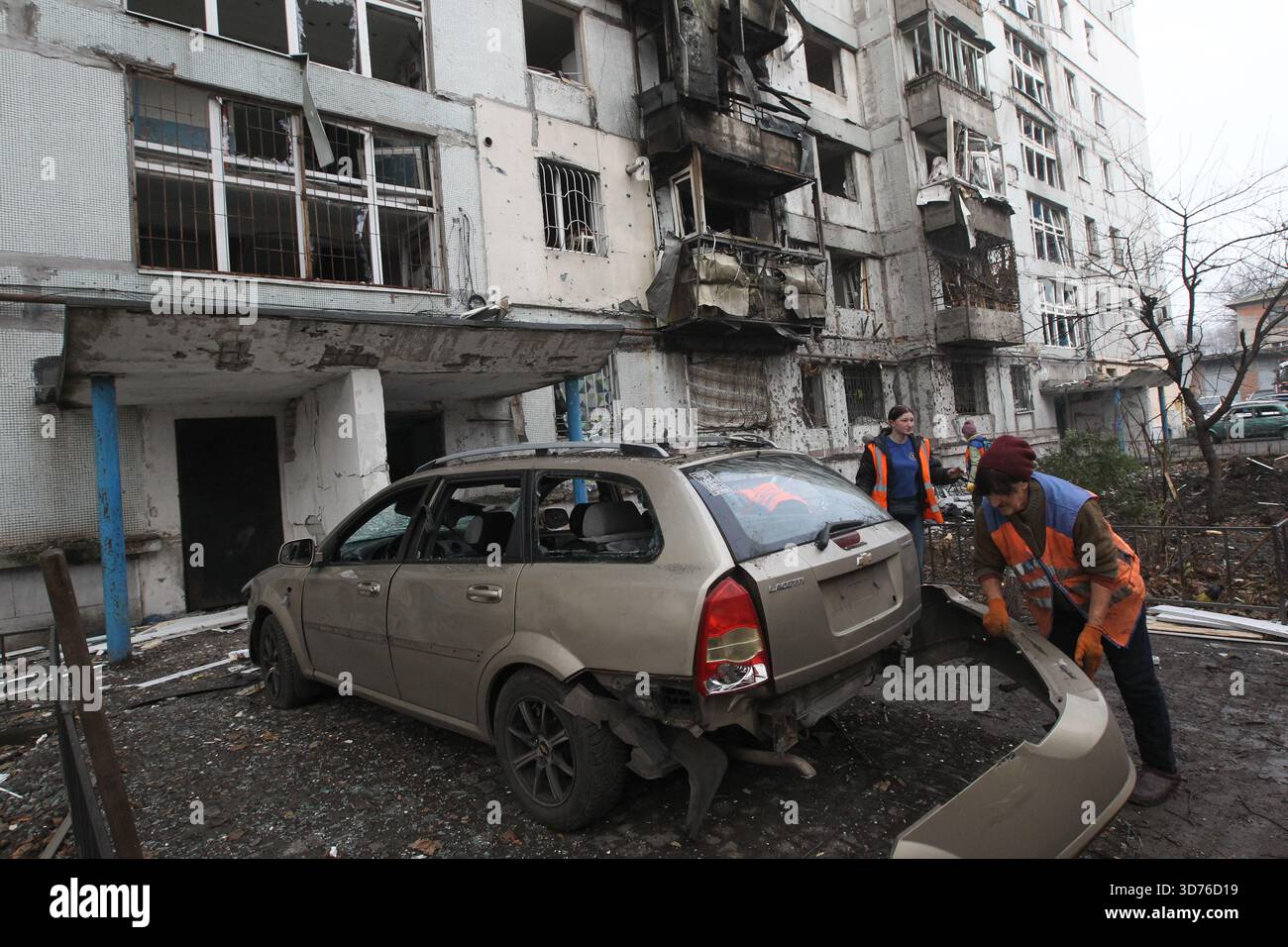 Ein Versorgungsarbeiter hält den hinteren Stoßfänger eines Autos vor einem neunstöckigen Wohnblock, der durch einen russischen Drohnenangriff beschädigt wurde, Dnipro, Ukraine, 23. November 2025. Über Nacht starteten russische Truppen einen großangelegten Angriff auf die Stadt, wobei sie Balkone eines neunstöckigen Wohngebäudes entzündeten und die Räumlichkeiten eines Privathauses beschädigten. Sechs Wagen wurden beim Streik beschädigt. 15 Menschen benötigten medizinische Hilfe. Drei sind noch immer stationär: Frauen im Alter von 52 und 64 Jahren und ein 48-jähriger Mann. Einer leidet an Vergiftungen durch Verbrennungsprodukte, während die anderen Kopfverletzungen und Verletzungen erlitten Stockfoto