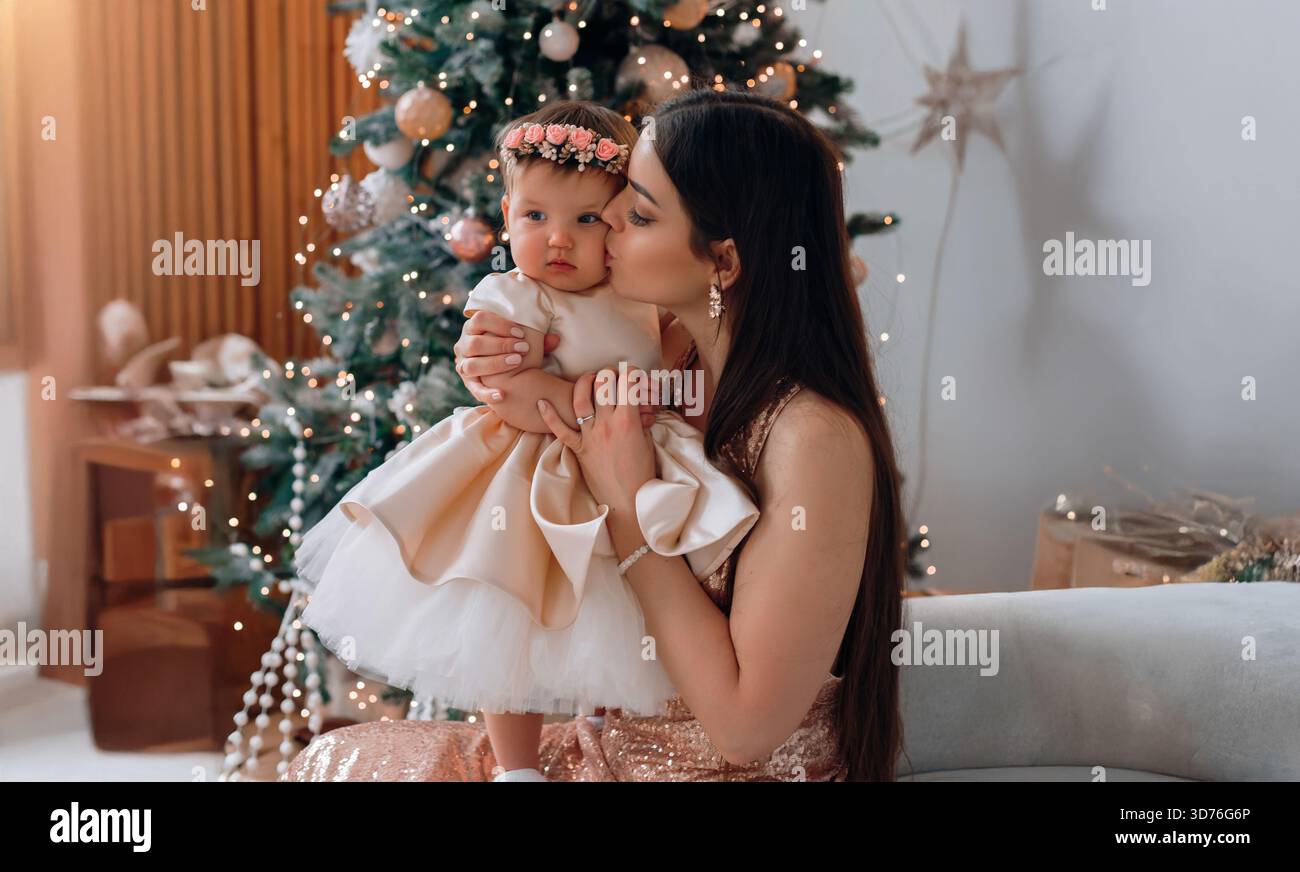 Ein freudiger Moment wird festgehalten, als eine Mutter ihr kleines Mädchen, das in einem wunderschönen Outfit gekleidet ist, vor einem glitzernden Weihnachtsbaum liebevoll küsst. Stockfoto