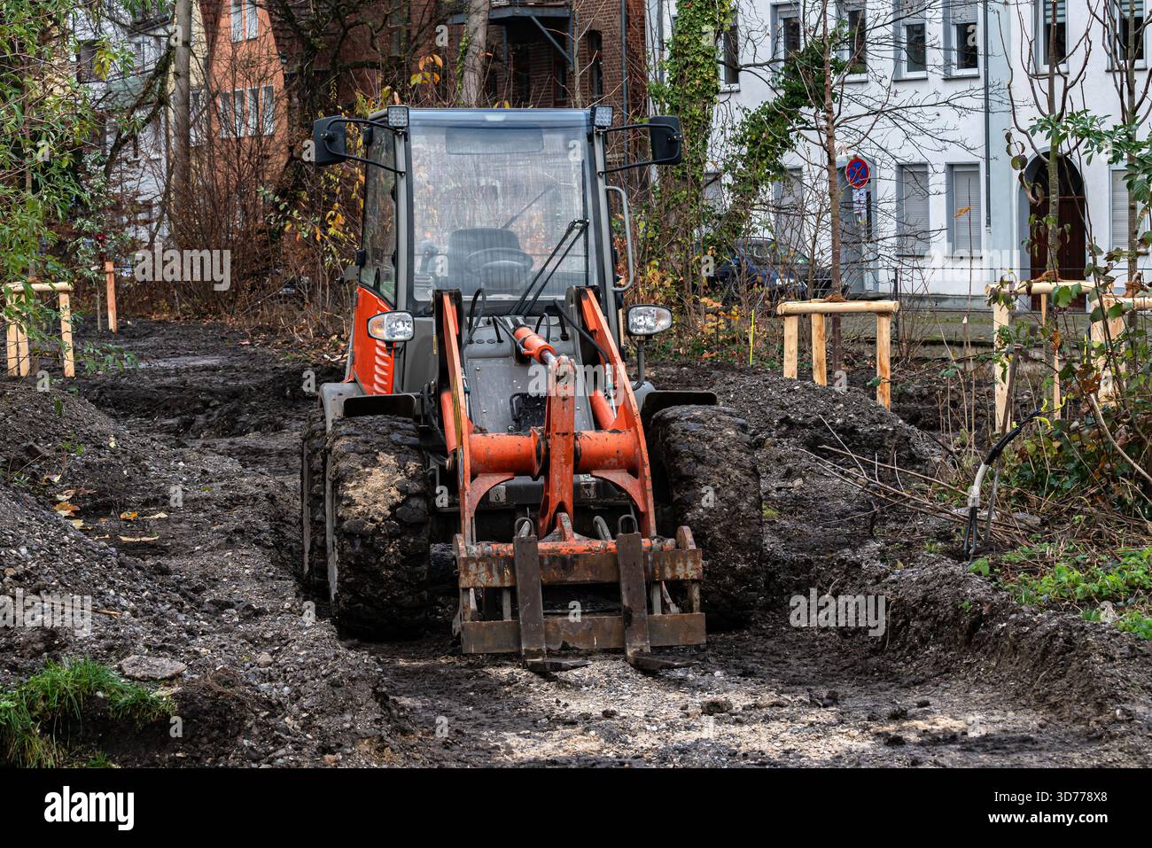 Ein Kompakttraktor arbeitet aktiv auf einer Baustelle, die von Gebäuden umgeben ist. Der Boden ist matschig und uneben, während die Maschine den Bereich vorbereitet Stockfoto