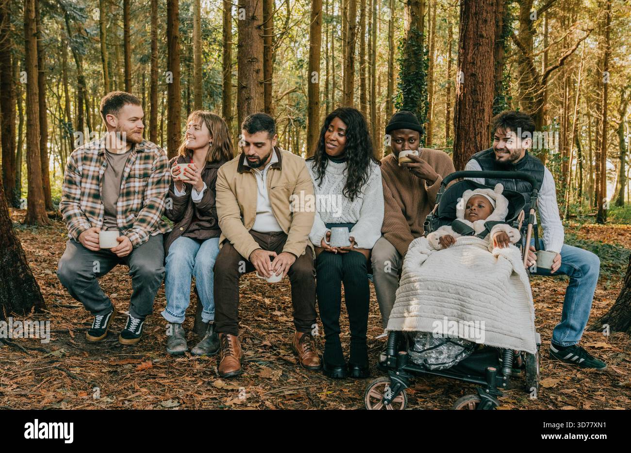 Eine Gruppe von Menschen sitzt in einem Wald, einer von ihnen schiebt einen Kinderwagen. Die Gruppe ist vielfältig, mit Menschen unterschiedlichen Alters und Rassen. Das sind sie Stockfoto