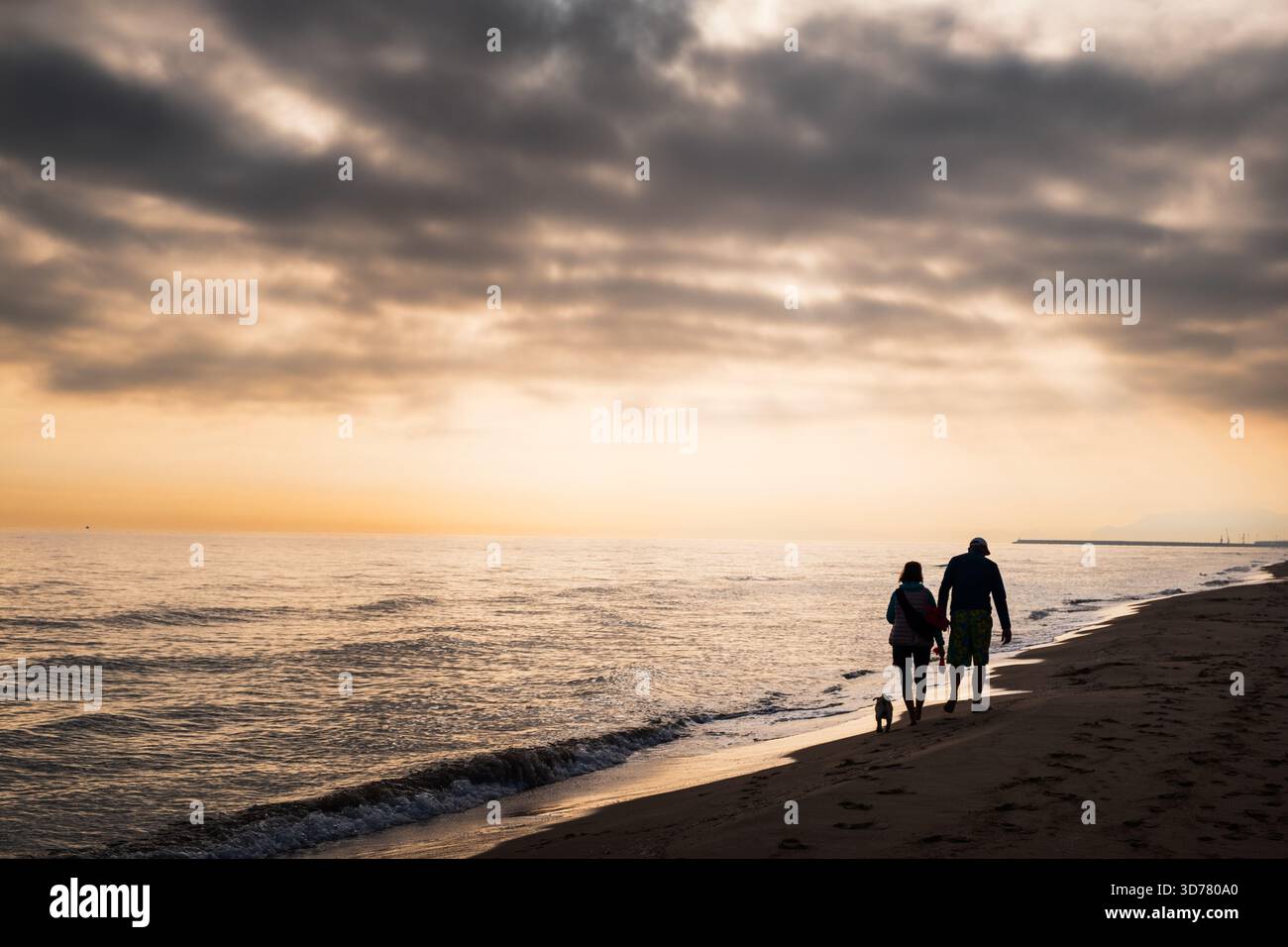 Ein paar Spaziergänge am Gandia Sandstrand in der Abenddämmerung, begleitet von einem verspielten kleinen Hund. Sanftes Licht der untergehenden Sonne reflektiert auf sanften Wellen Stockfoto