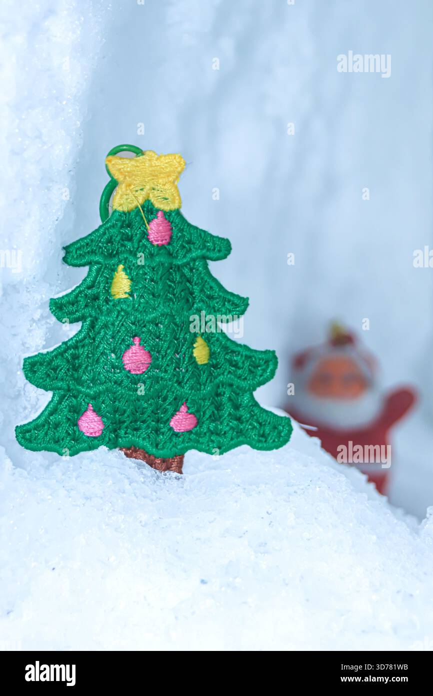 Gestickter Weihnachtsbaum auf Gletscherschnee mit einem entfernten verspielten Weihnachtsmann in sanfter Unschärfe. Eine skurrile Winter-Mikrowelt, perfekt für Urlaubsthemen. Stockfoto