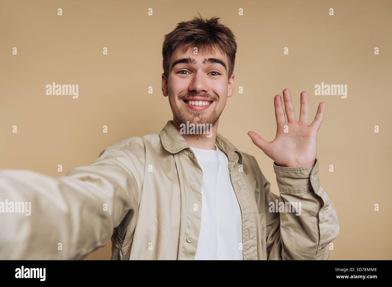 Ein junger Mann lächelt und winkt mit der Hand, während er ein Selfie macht. Er trägt ein helles Hemd und hat kurze Haare, steht vor einem einfachen Beige Stockfoto