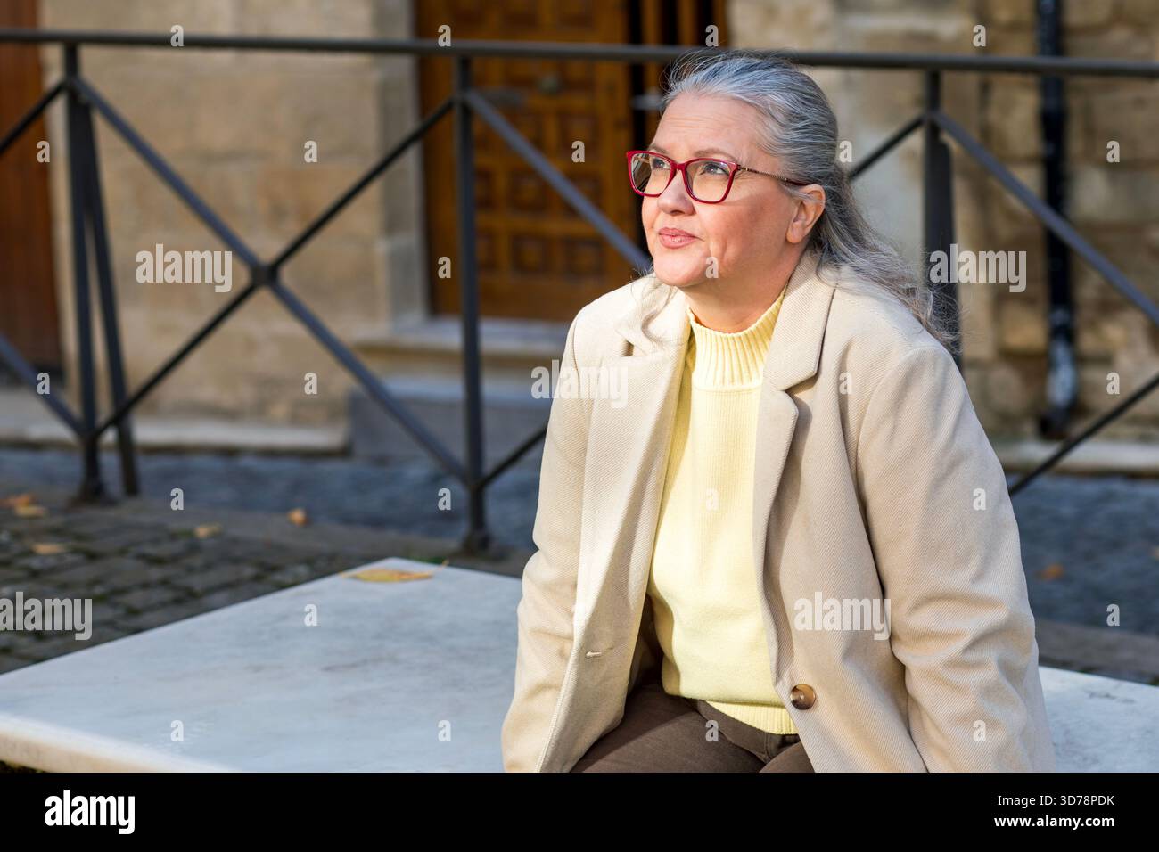 In einer malerischen, historischen Gegend sitzt eine Frau mit silbernen Haaren und Brillen auf einer Steinbank und trägt einen leichten Pullover und Blazer. Sie blickt nachdenklich, en Stockfoto