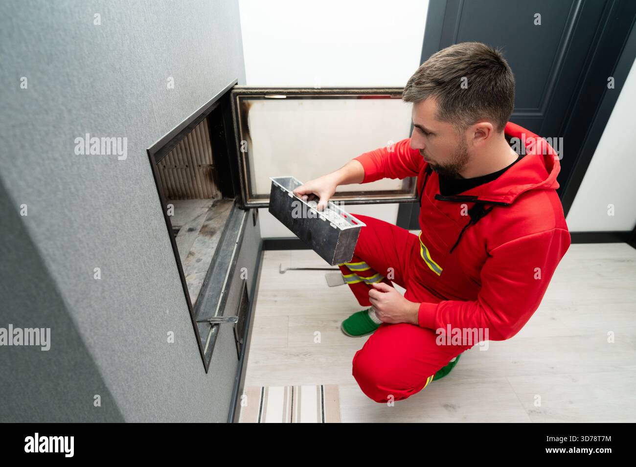 Ein Techniker in einem roten Jumpsuit inspiziert und reinigt einen eleganten Kamineinsatz in einem stilvollen Wohnzimmer sorgfältig und konzentriert sich dabei auf effiziente Funktionalität und Stockfoto