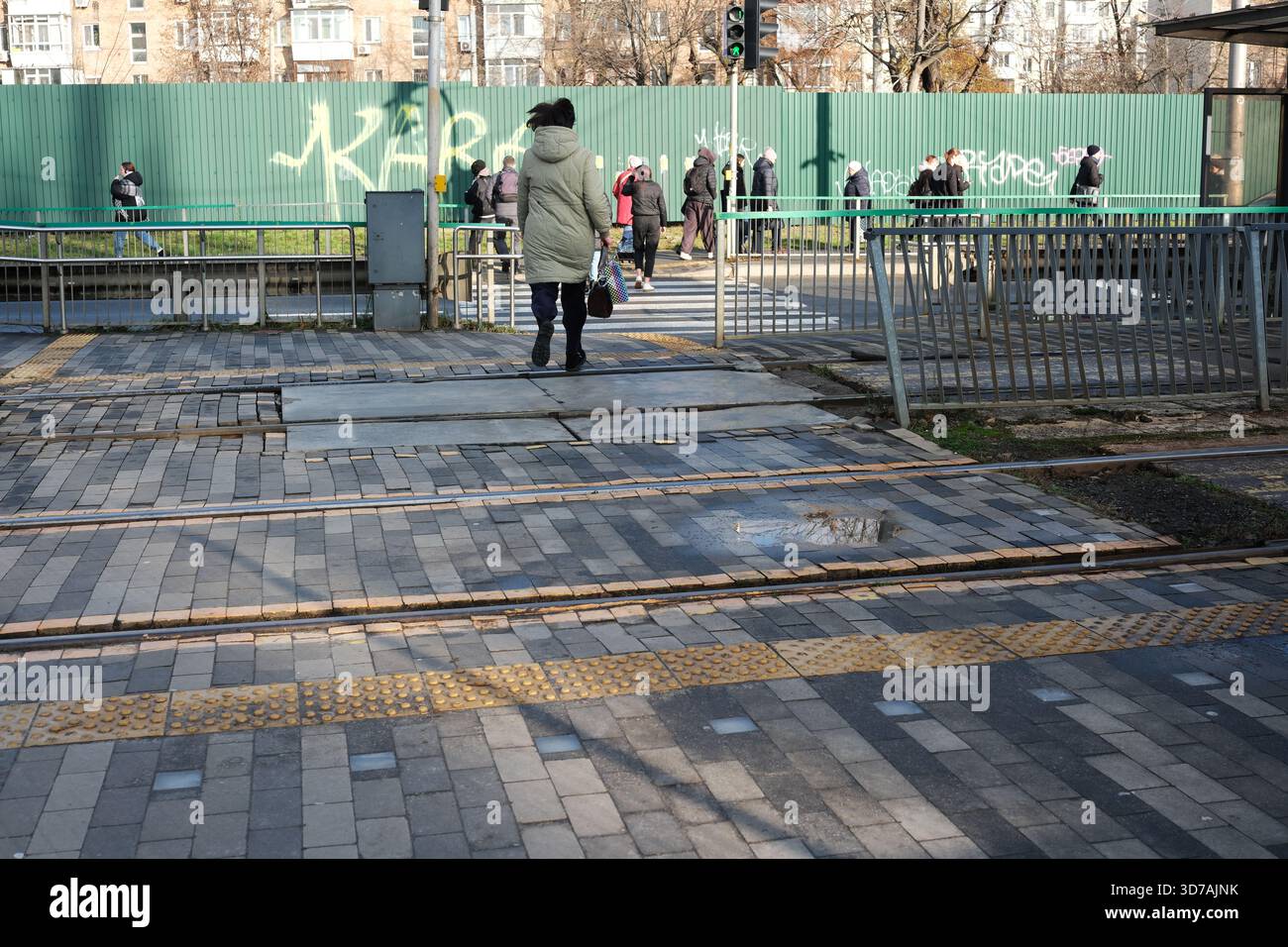 Fußgänger laufen durch eine lebhafte Stadtüberquerung mit Bahngleisen unter Sonnenlicht. November 2025 Kiew, Ukraine Stockfoto