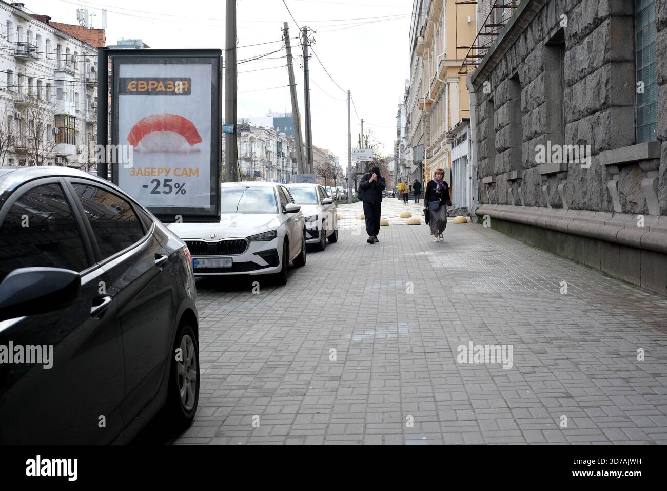 Zwei Personen spazieren auf einem sauberen Bürgersteig, der von geparkten Autos gesäumt ist. November 2025 Kiew, Ukraine Stockfoto