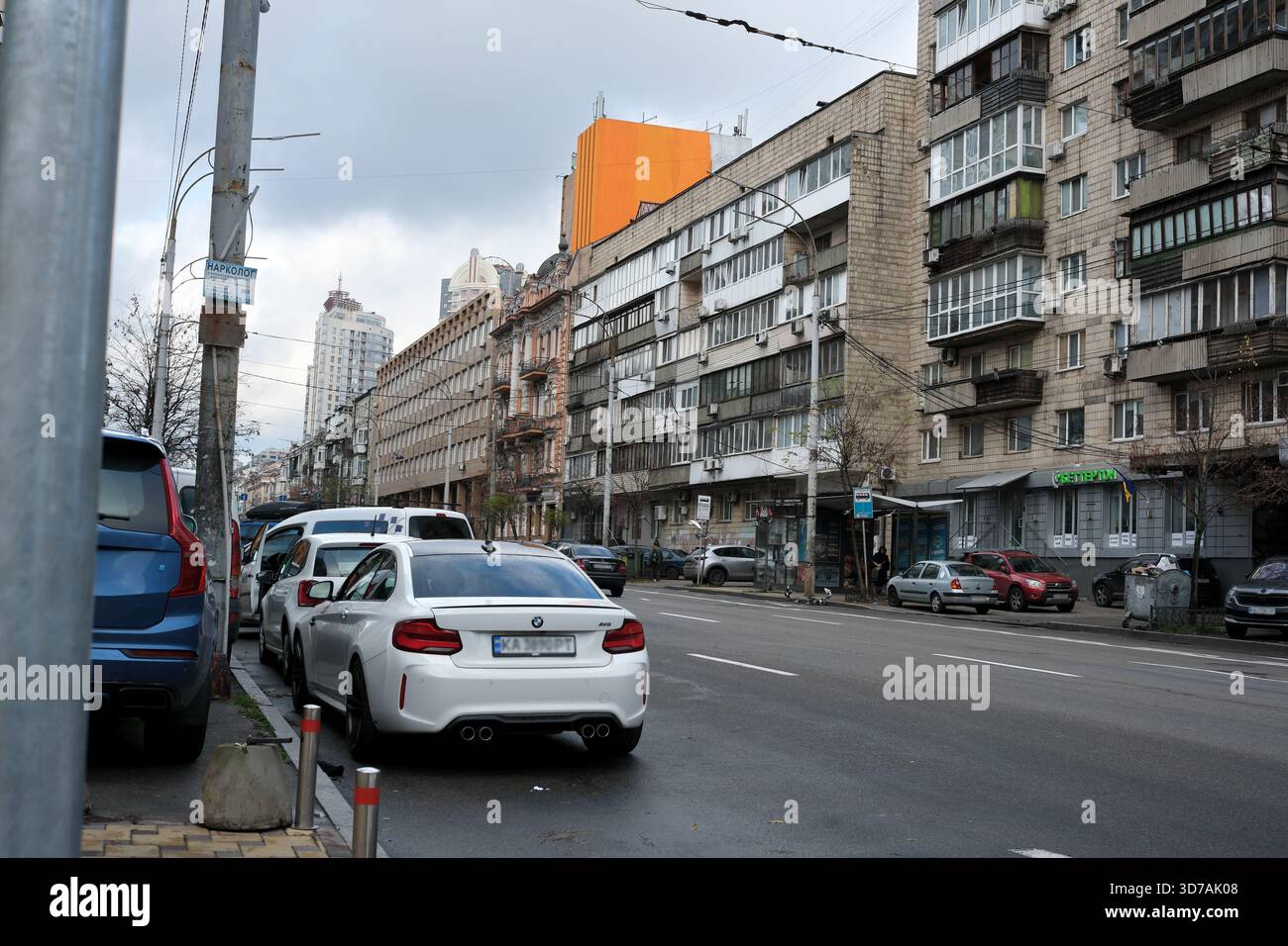 Autos säumen die Straße, während sich hohe Gebäude gegen einen bewölkten Himmel im Stadtbild erheben. November 2025 Kiew, Ukraine Stockfoto