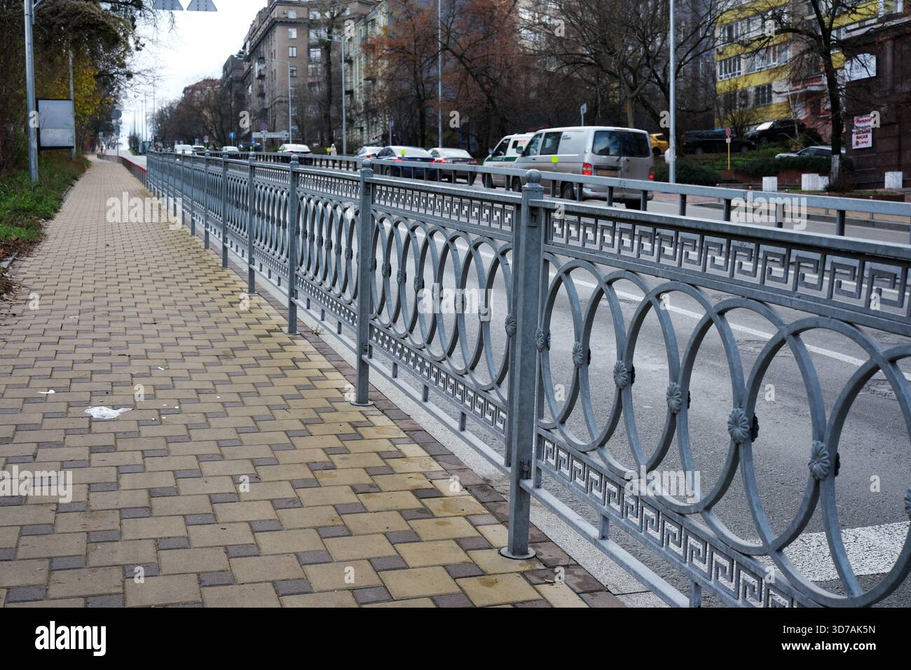 Ein dekorativer Metallzaun grenzt an einen ruhigen Bürgersteig neben einer belebten Straße. November 2025 Kiew, Ukraine Stockfoto