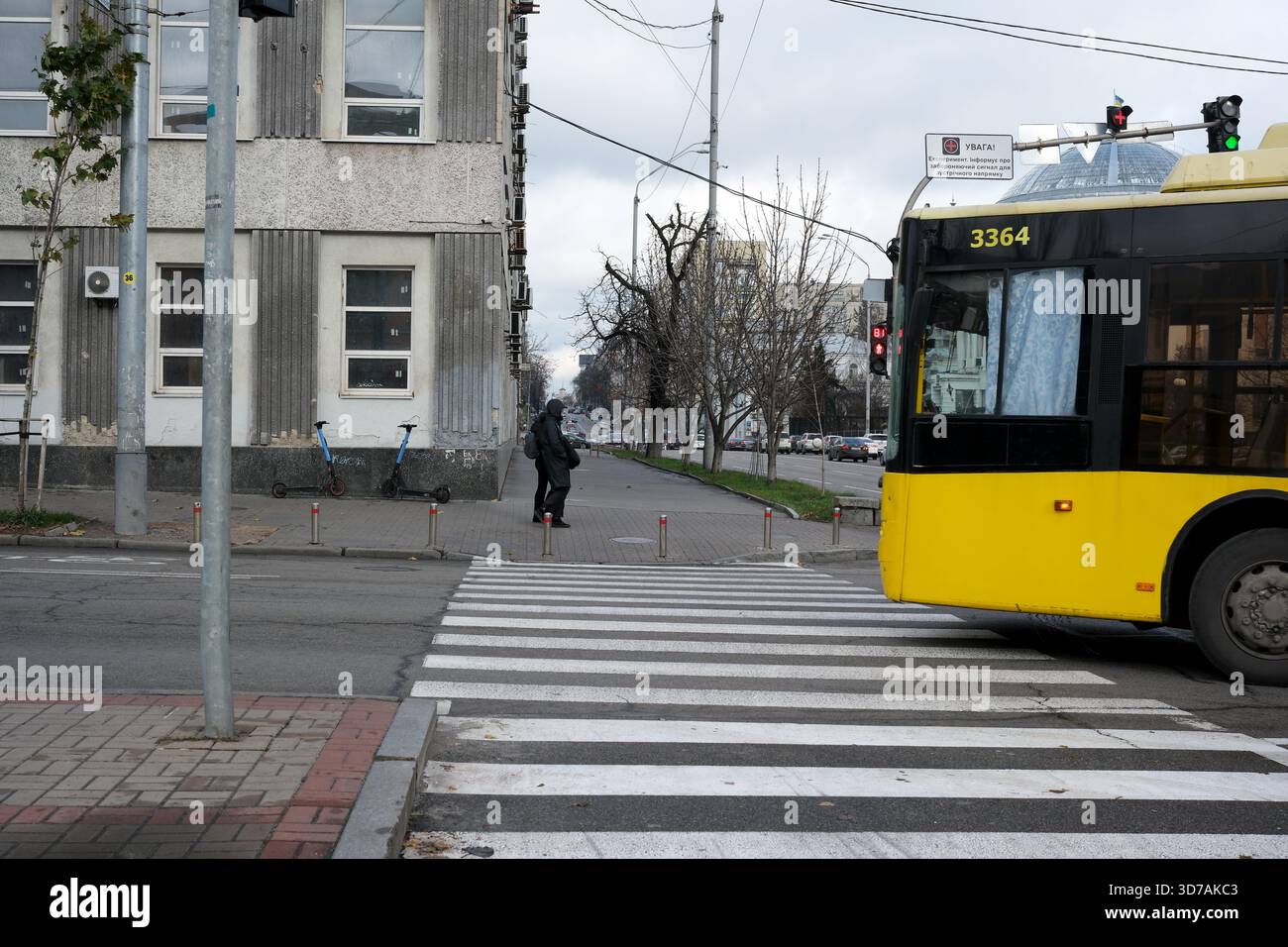 Ein gelber Bus hält in der Nähe eines Fußgängers, der am Straßenübergang unter grauem Himmel wartet. 24. November 2025, Kiew, Ukraine, Schewtschenko Boulevard Stockfoto
