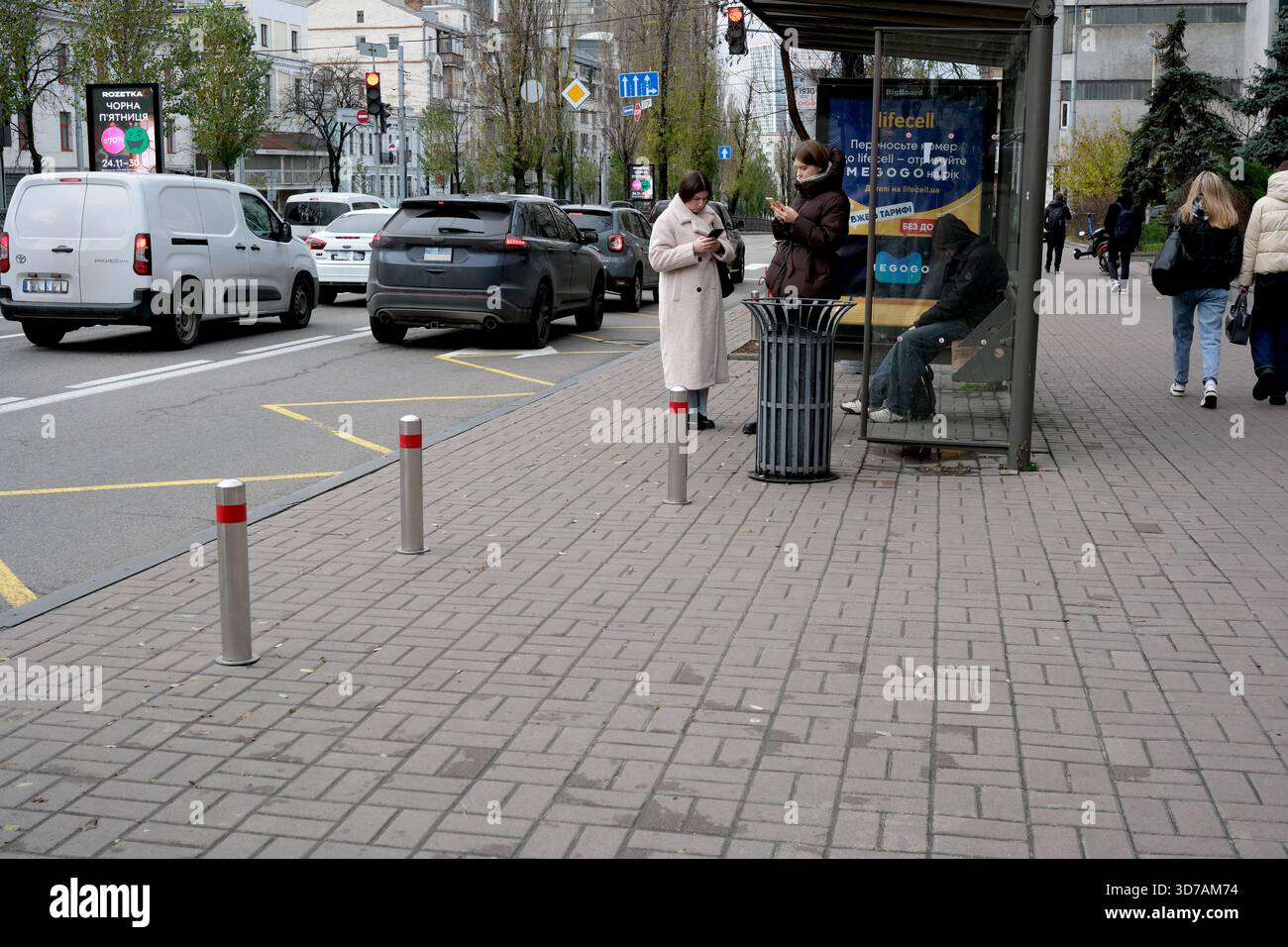 Zwei Frauen stehen an einer Bushaltestelle, während andere auf einer belebten Stadtstraße vorbeilaufen. 24. November 2025, Kiew, Ukraine, Schewtschenko Boulevard Stockfoto