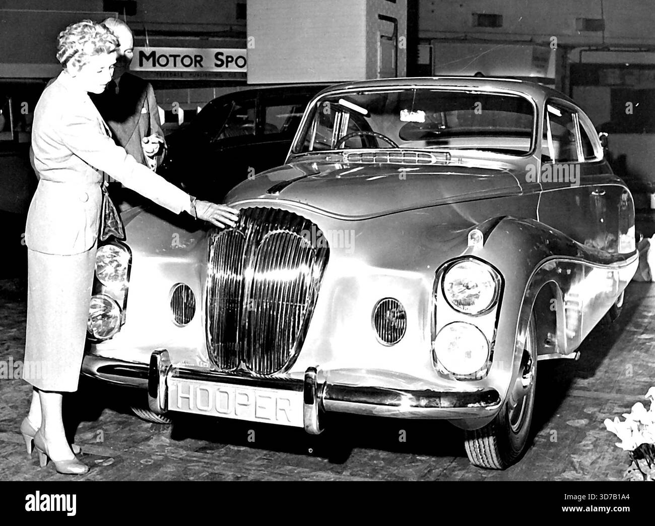 Lady Docker, die fabelhafte Lady Docker, hat ein neues Auto. Im Vergleich zu ihrem letzten Daimler ist es eine ziemlich einfache kleine Angelegenheit, mit dem echten goldenen Heizkörper und den Armaturen, die überall für Furore sorgen. Nur ein Abstecher für ihren persönlichen Gebrauch, dieses neue Auto, versichert Lady Docker. Der Docker Silver Flash Daimler hat die Vorschau geklaut. In Kombination mit der Beredsamkeit von Lady Docker, die jeden der Verkäufer auf jedem Stand überbot, indem sie die Tugenden ihres Runabout lobte, zog es die Massen wie Stahl zu einem Magneten. Ihr Ehemann, Sir Bernard Docket, machte die Aufmerksamkeit auf das Armaturenbrett, die Türverkleidungen und die AR Stockfoto