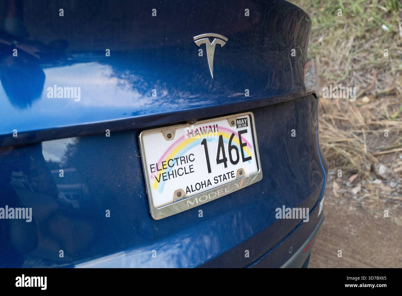 Nahaufnahme eines hawaiianischen Kennzeichenschilds mit dem Regenbogenmuster „Aloha State“ und der Bezeichnung „Elektrofahrzeug“ auf einem blauen Tesla Model Y Stockfoto