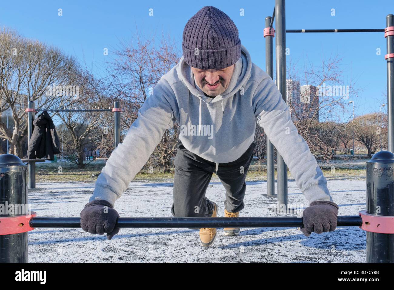 Reife Mann konzentriert sich intensiv auf Liegestütze an parallelen Bars im Outdoor Calisthenics Gym. Das Training findet an kalten, sonnigen Tagen mit Schnee statt Stockfoto