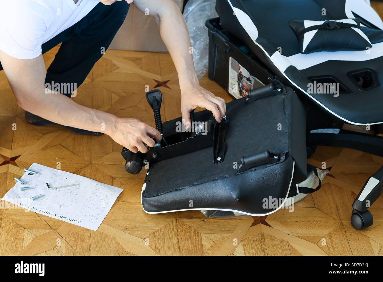 Ein Mann in weißem Hemd kniet auf Holzboden und befestigt den schwarz-weißen Sitzplatz des Spielstuhls mit Befestigungsteilen an seiner Basis. Stockfoto