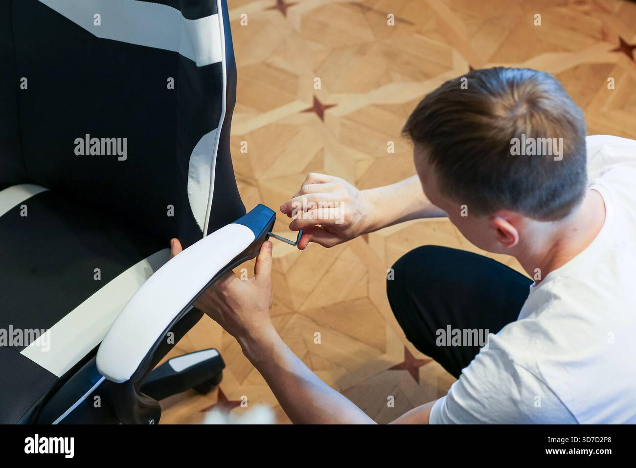 Hände einer Person, die die Armlehne eines schwarz-weißen ergonomischen Spielstuhls mit einem Schraubendreher auf einem Holzboden mit Sternenmuster sichern. Stockfoto