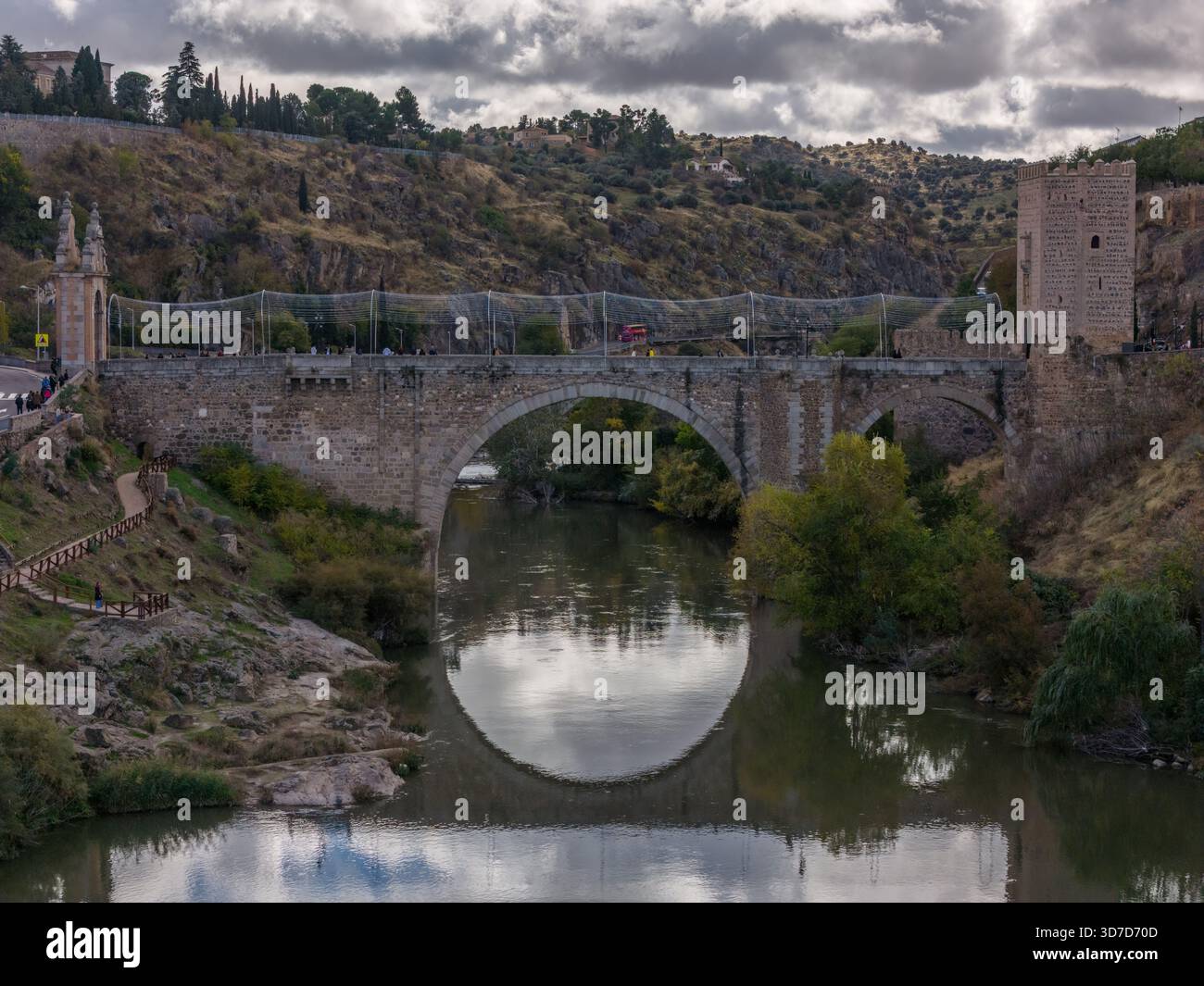 Aus der Vogelperspektive der alten Alcantara-Brücke erhebt sich anmutig über den ruhigen Fluss Tejo unter einem bewölkten Himmel, Toledo, Kastilien-La Mancha, Spanien. Stockfoto