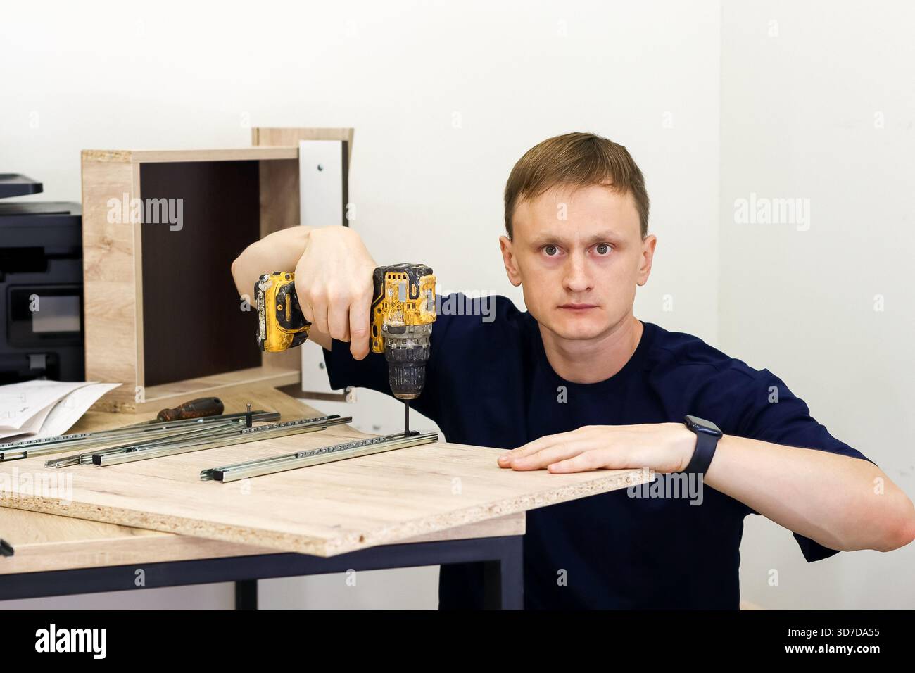 Zimmermann mit Bohrmaschine zum Befestigen des Schubladenschiebers auf Holzbrett. Zimmermann in dunklem Hemd mit Bohrmaschine zum Befestigen der Metallschublade auf Holz b Stockfoto