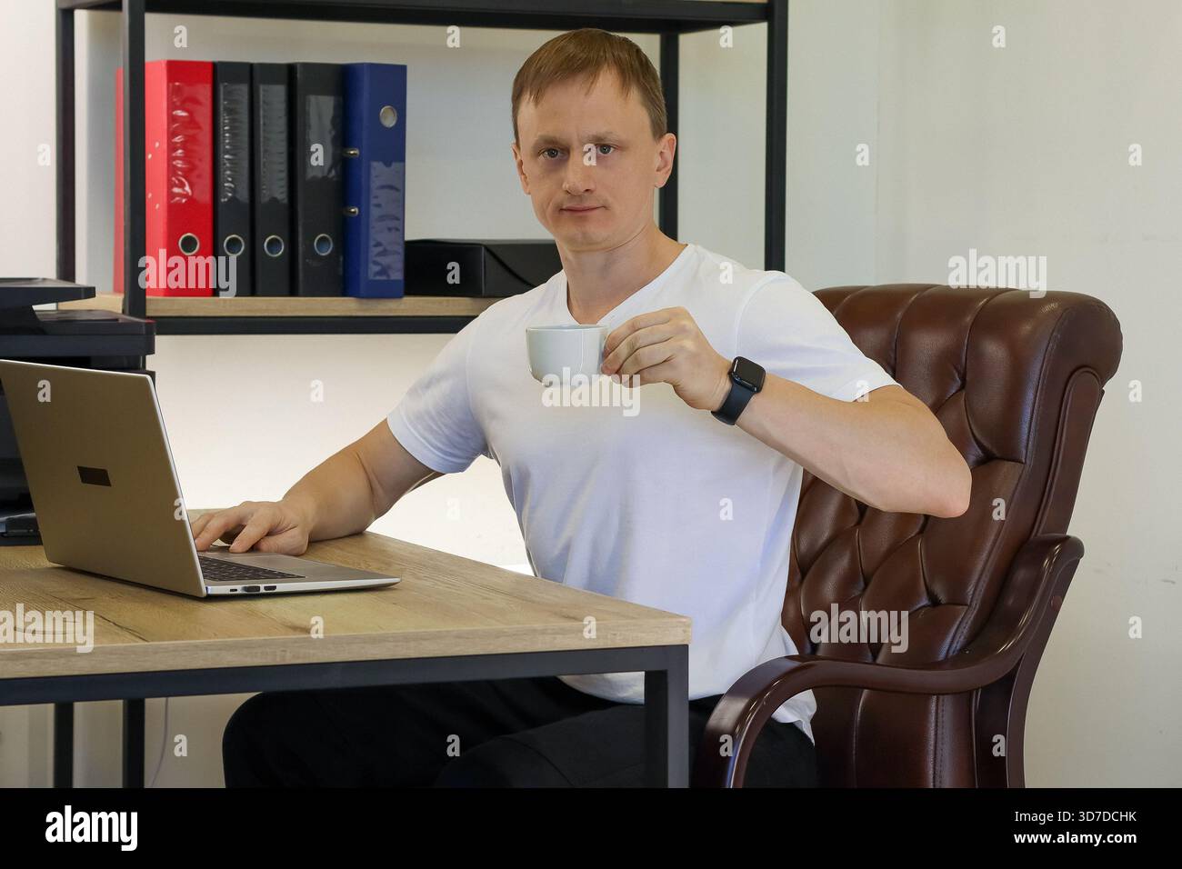 Junger Mann in weißem T-Shirt, der einen weißen Becher hält, während er am Laptop am Holztisch im Büro arbeitet Stockfoto
