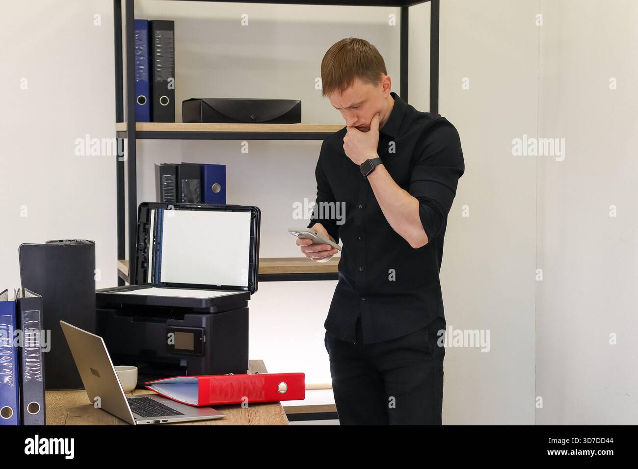 Ein durchdachter Geschäftsmann berät ein mobiles Gerät neben einem fehlerhaften Drucker im Arbeitsbereich Stockfoto
