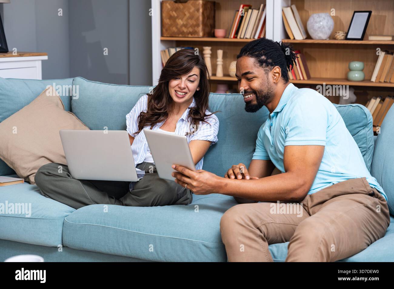 Ein lächelndes Paar sitzt auf einem Sofa und vergleicht ein Tablet mit einem Laptop, koordiniert Aufgaben für Budgetierung, Studium oder ein kleines Geschäftsprojekt. Echtes Zuhause kollabora Stockfoto