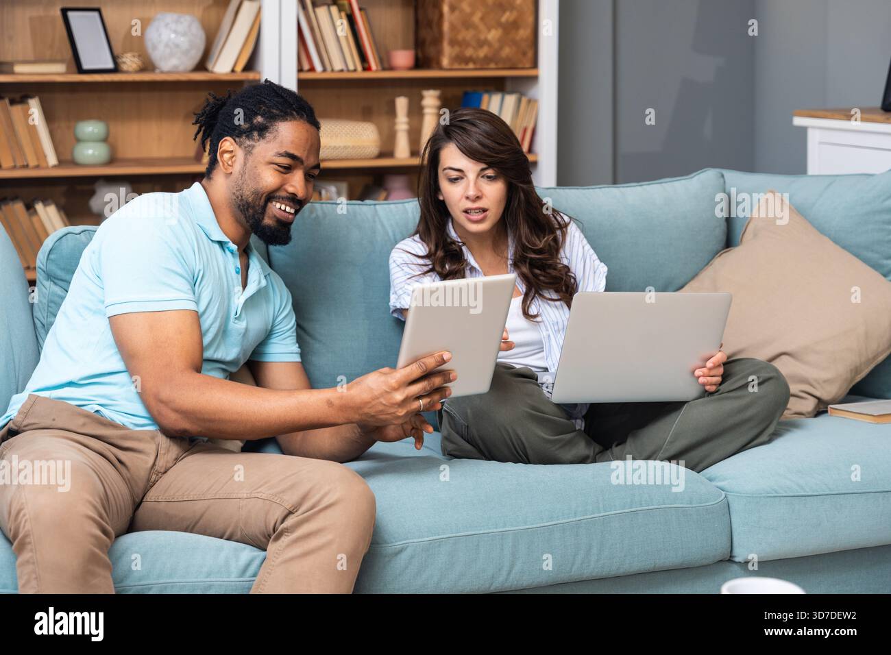Ein lächelndes Paar sitzt auf einem Sofa und vergleicht ein Tablet mit einem Laptop, koordiniert Aufgaben für Budgetierung, Studium oder ein kleines Geschäftsprojekt. Echtes Zuhause kollabora Stockfoto