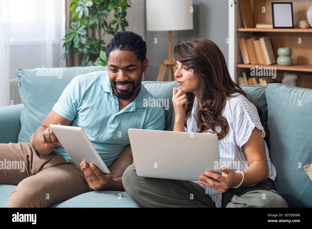 Ein lächelndes Paar sitzt auf einem Sofa und vergleicht ein Tablet mit einem Laptop, koordiniert Aufgaben für Budgetierung, Studium oder ein kleines Geschäftsprojekt. Echtes Zuhause kollabora Stockfoto
