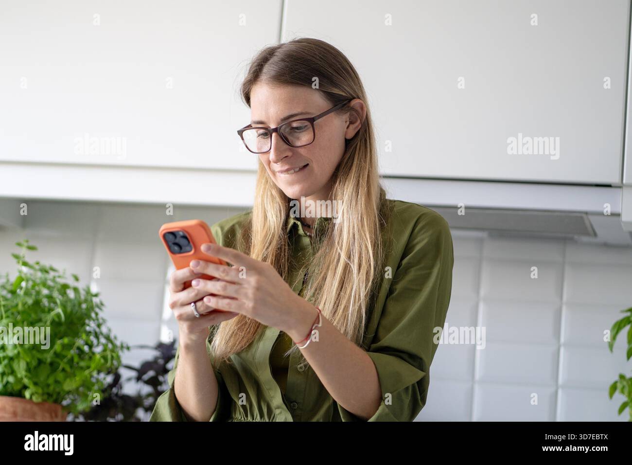 Positive Frau, die Nachrichten auf dem Smartphone schreibt, während sie in der Küche zu Hause mit einem Lächeln steht. Online-Kommunikation per Telefon, Chat und Online-Shopping, Webfeed für Frauen und soziale Medien zu Hause || Model veröffentlicht Stockfoto