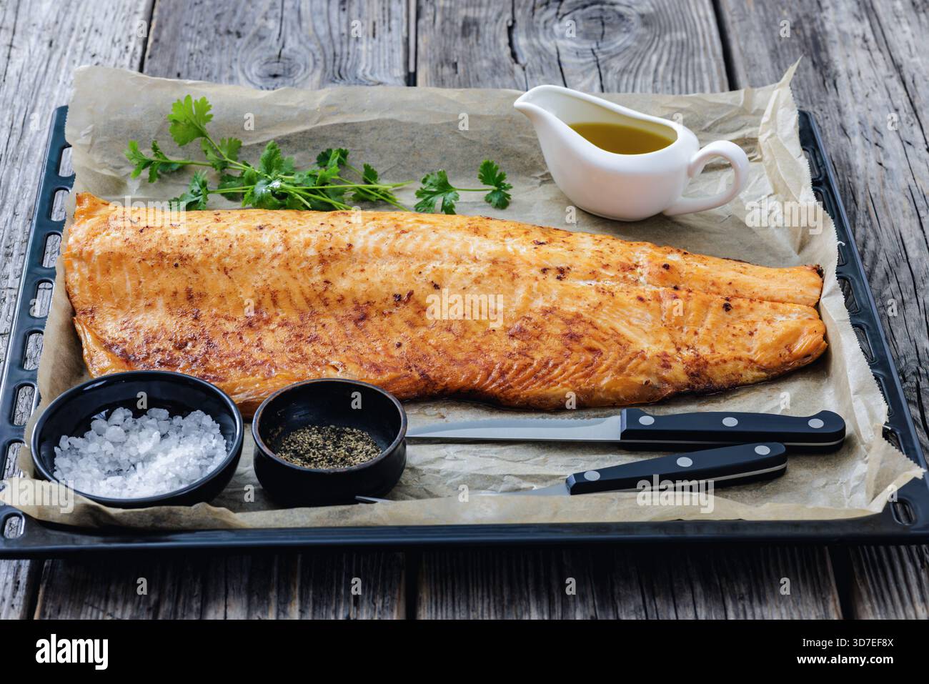 Gebratenes Lachsfilet auf Backpapier auf Backblech mit Salz, Pfeffer, Besteck, auf rustikalem Holztisch, horizontale Sicht von oben, nicht AI ge Stockfoto