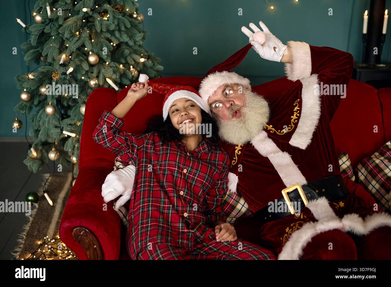 afroamerikanisches Mädchen im roten Pyjama, das Selfie mit dem weihnachtsmann auf roter Couch macht Stockfoto