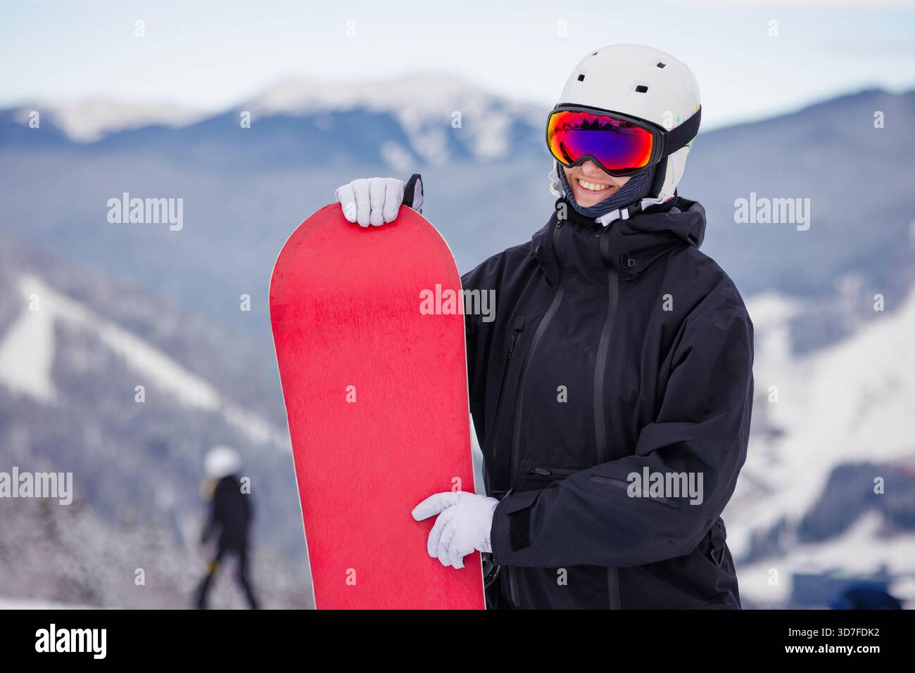 Fröhlicher junger Snowboarder in schwarzer Jacke und Helm mit hellrotem Brett auf verschneiten Berghängen Wintersporturlaubskonzept. Stockfoto