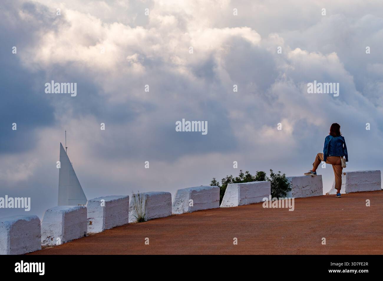 Eine dramatische Komposition mit einer einsamen Figur, die auf einer weißen Barrikade steht und über eine bewölkte, atmosphärische Szene blickt. Eine weiße, segelähnliche Struktur Stockfoto