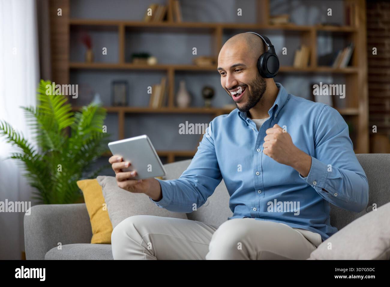 Ein Mann mit Kopfhörern auf der Couch feiert den Sieg, während er ein Tablet ansieht und seine Faust bei entspanntem Heimspiel, Streaming oder Videoanruf-Momenten mit Freude zusammendrückt Stockfoto