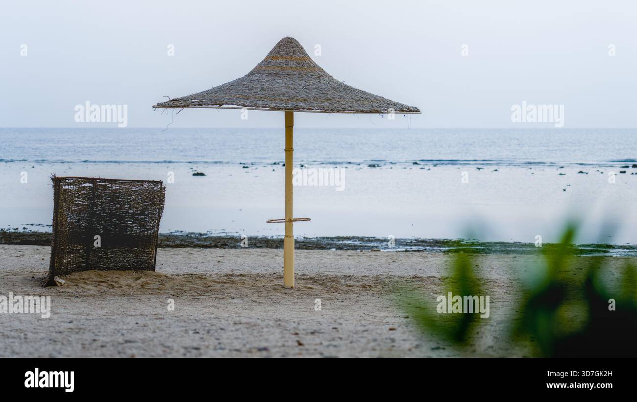 Ein einzelner Sonnenschirm und Windschutz stehen ruhig am Strand eines ägyptischen Resorts im frühen Morgenlicht. Stockfoto