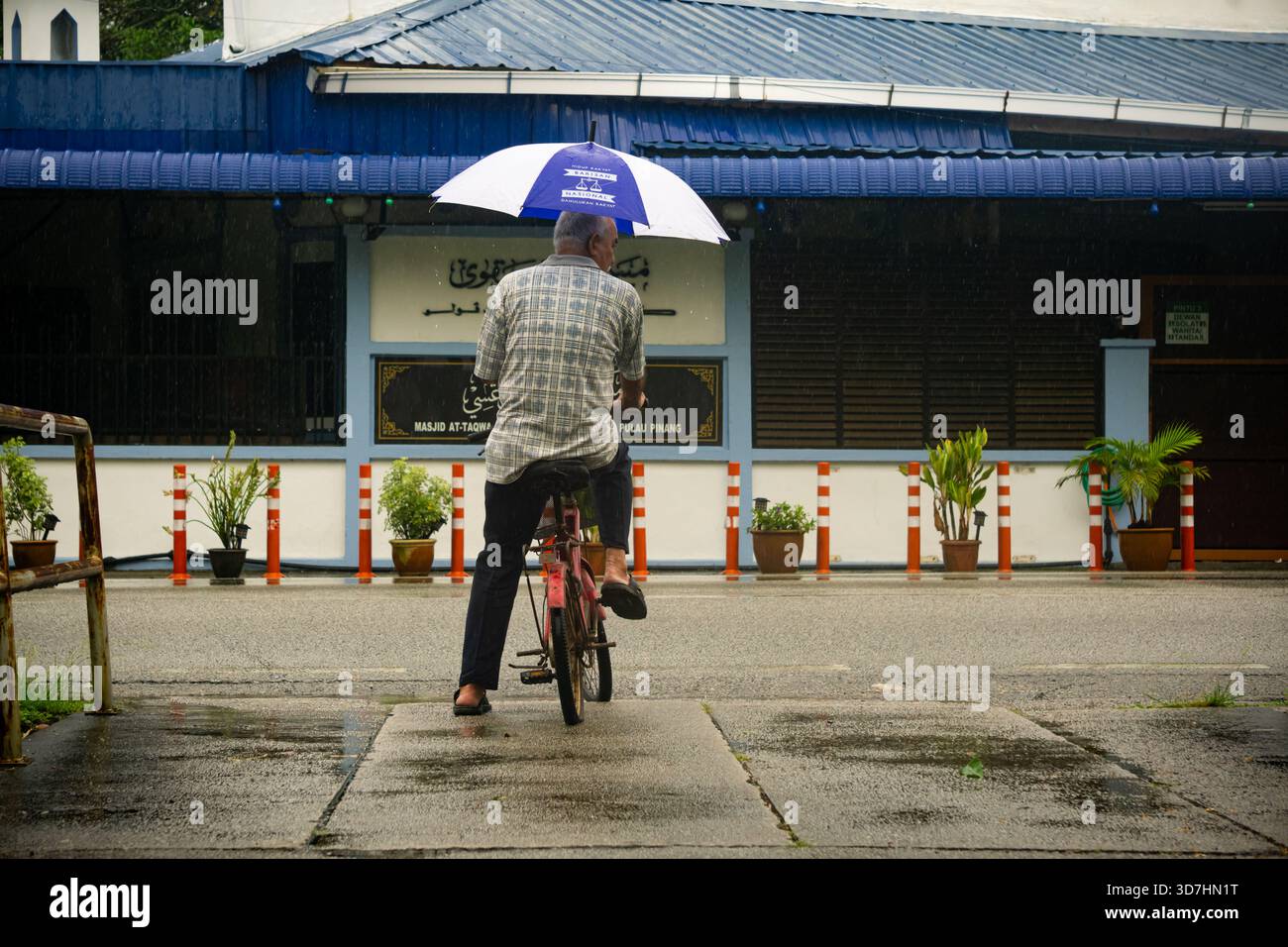 Ein älterer Mann radelt unter einem Regenschirm im Regen vor Masjid at-Taqwa, Balik Pulau, Penang. Ein ruhiger Moment des täglichen Lebens in einem malaysischen Dorf Stockfoto