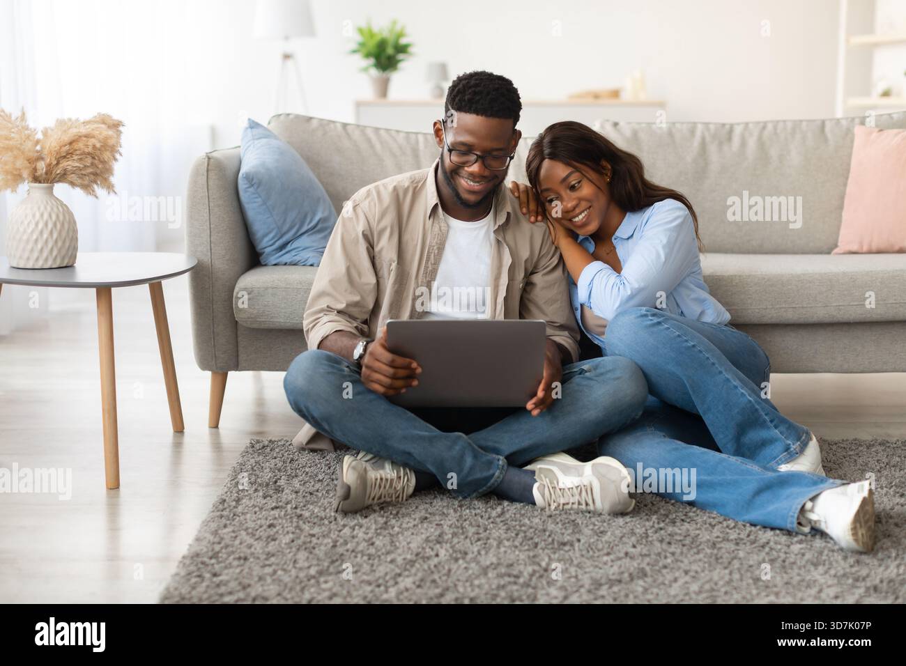 Junges schwarzes Paar genießt die gemeinsame Zeit, während es im gemütlichen Wohnzimmer mit einem Laptop arbeitet Stockfoto