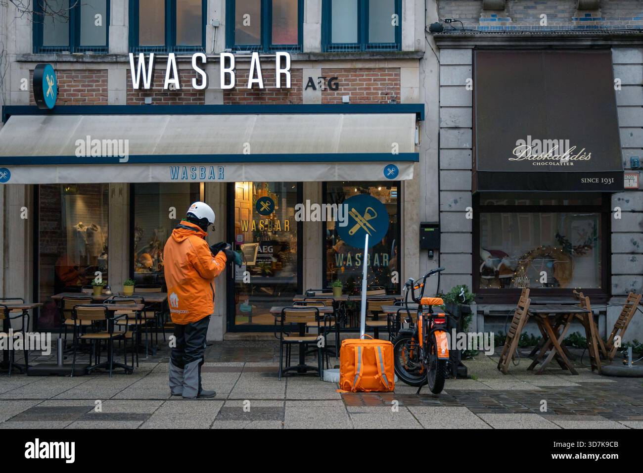 Fahrer mit Essenslieferung in einer orangefarbenen Jacke vor einem Wasbar Restaurant im Zentrum von Gent. Stockfoto
