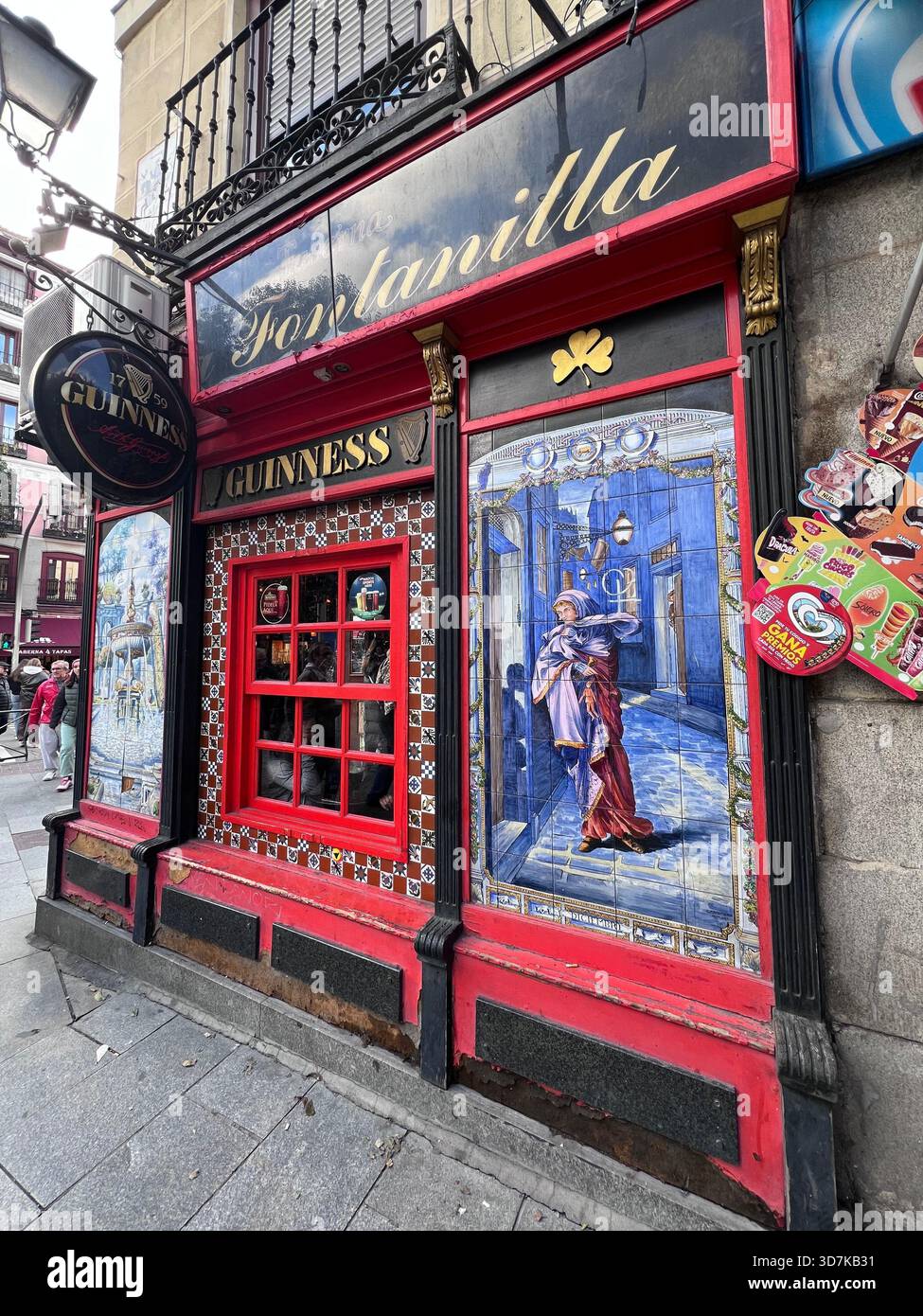 Traditionelle Pub-Fassade in Madrid mit rotem Fensterrahmen, Guinness-Schild und farbenfrohem Keramikfliesen an einer belebten Straße in Spanien. Stockfoto