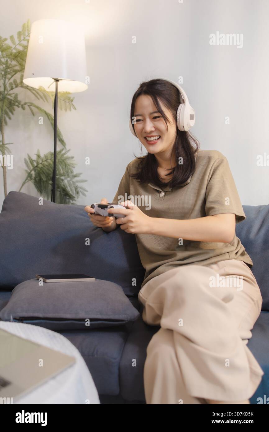 Eine junge Frau spielt zu Hause gern Videospiele, trägt Kopfhörer und hält einen Controller mit Spannung. Sie sitzt bequem auf einem Sofa und lächelt Stockfoto